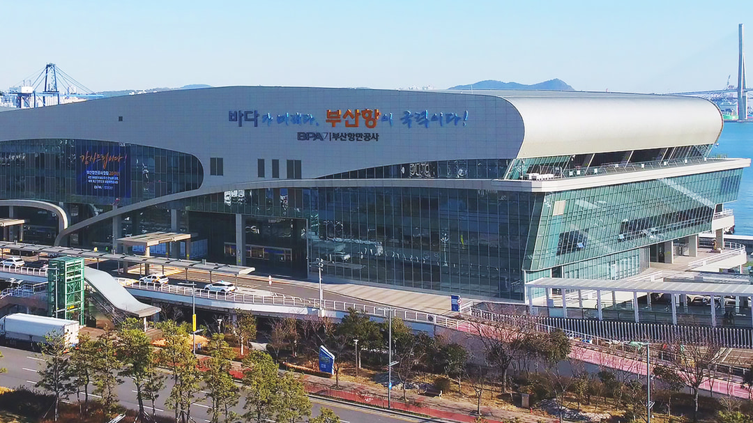부산항국제여객터미널 전경. 바다를 따라 길게 뻗은 곡선 지붕의 유리 건물이며, 정면 벽에 부산항만공사 표시가 있습니다. 오른쪽 멀리 부산항대교가 보입니다.